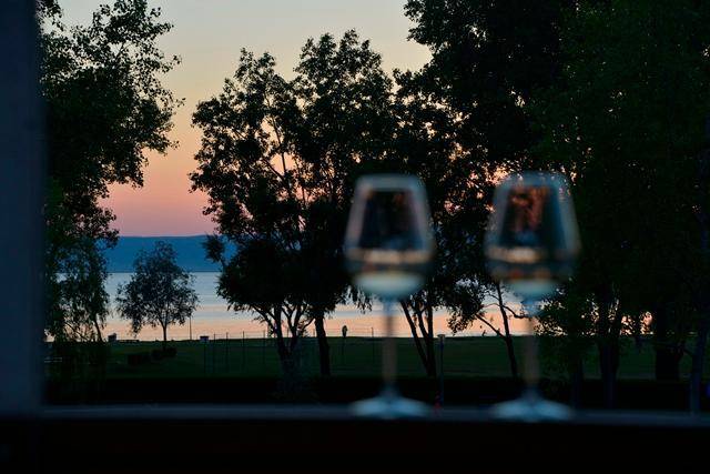 Abendlicher Blick auf den See durch ein Fenster, umrahmt von Bäumen und mit Weingläsern im Vordergrund.