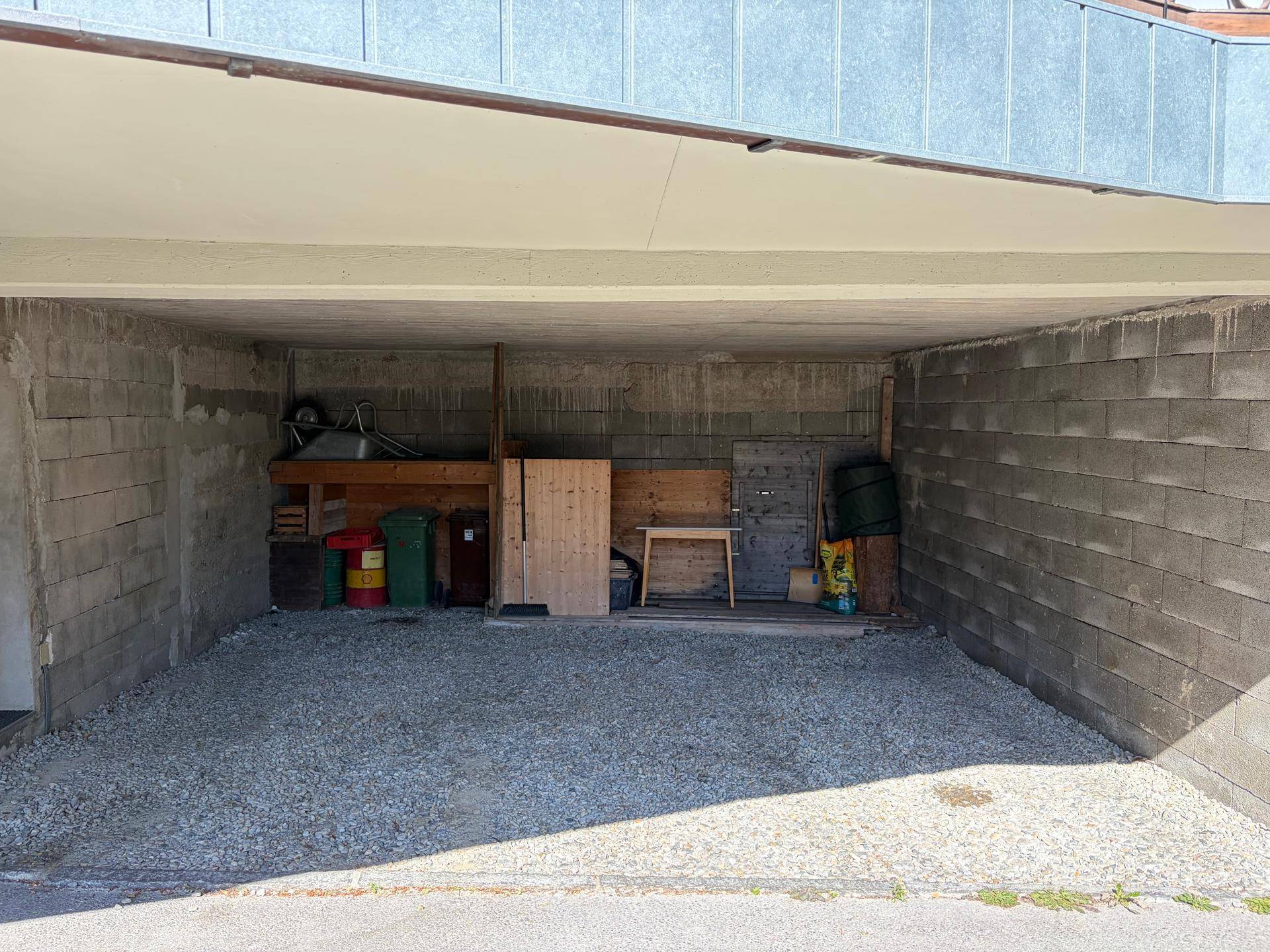 Offene Garage oder Carport mit Betonwänden und Kiesboden, genutzt als Abstellfläche.