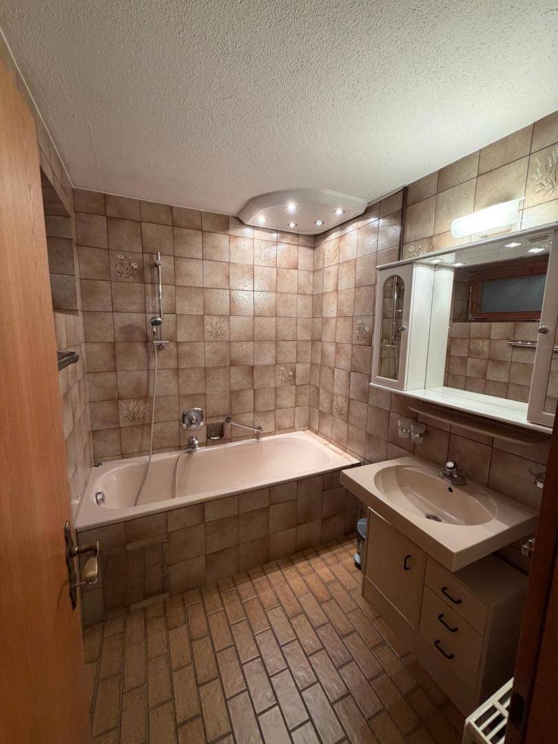 Badezimmer mit braunen Fliesen, Badewanne mit Duschfunktion und Spiegelschrank.