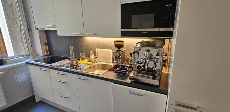 Kompakte Küchenzeile mit weißen Schränken, Spüle, Mikrowelle und einer professionellen Kaffeemaschine.