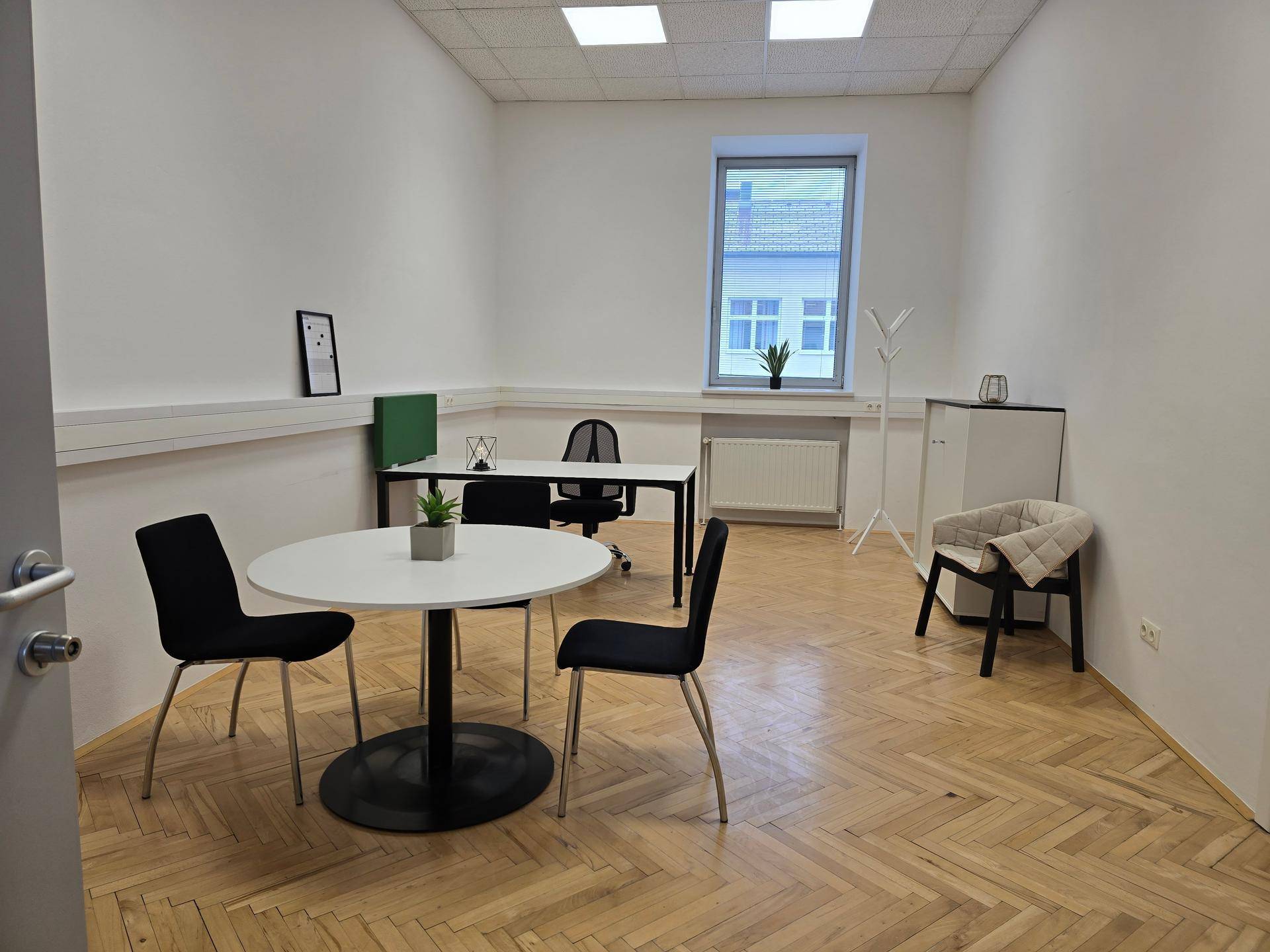Geräumiges Büro mit Fischgrätparkett, Besprechungstisch und Arbeitsplatz am Fenster.
