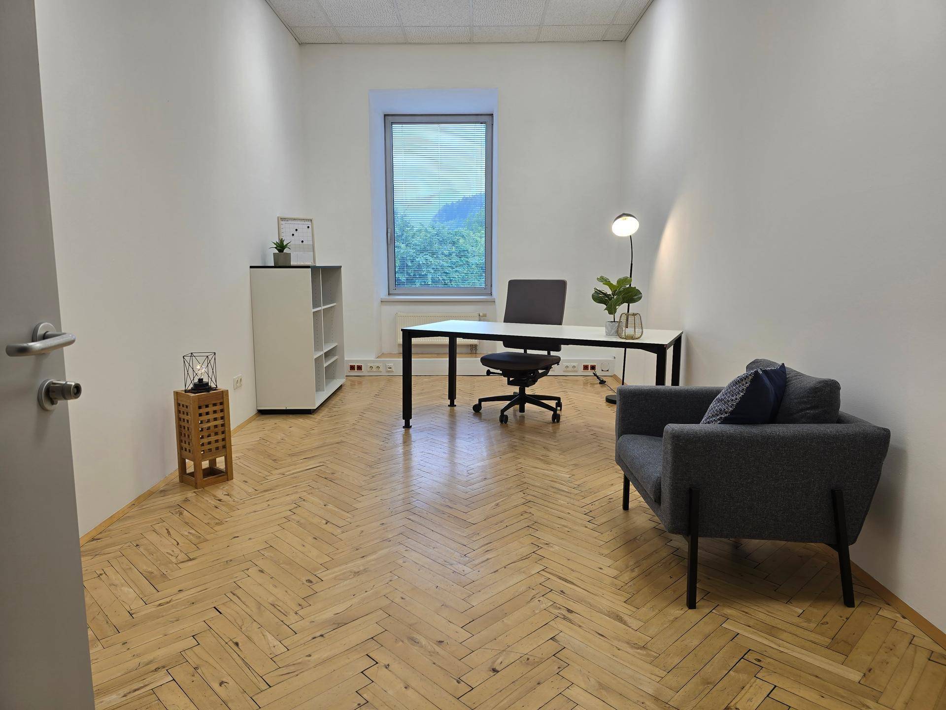 Helles Büro mit Fischgrätparkett, modernem Schreibtisch, Sessel und Blick ins Grüne.