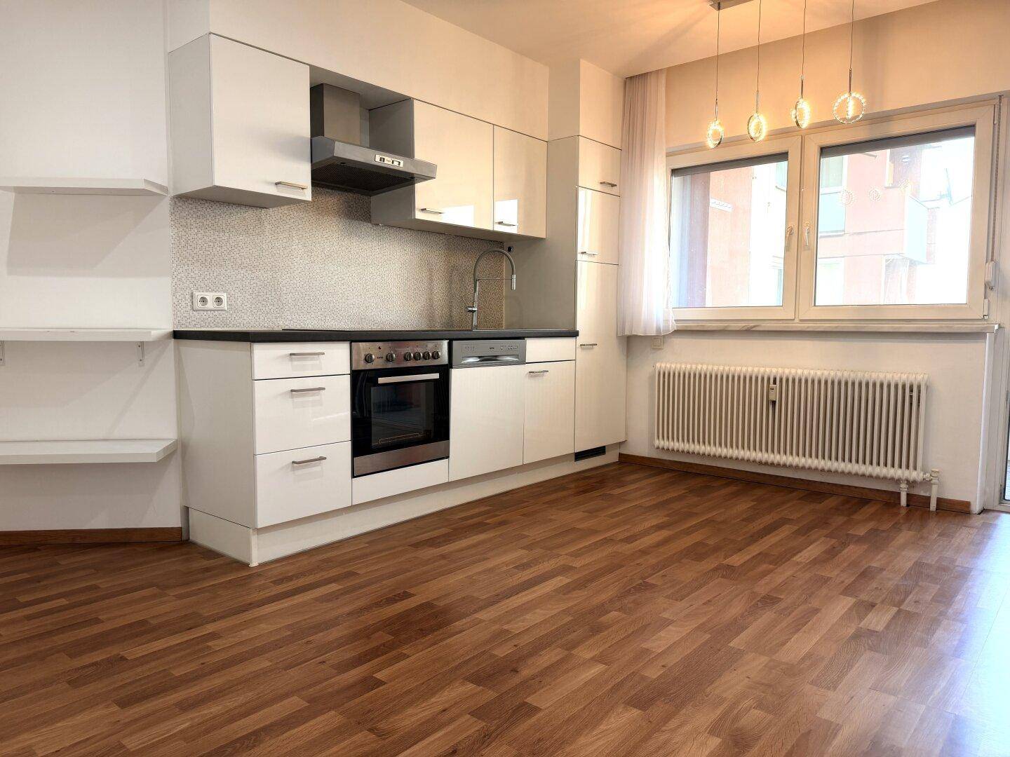 Moderne Einbauküche mit weißen Hochglanzfronten, dunkler Arbeitsplatte und Holzoptikboden in einem hellen Raum.
