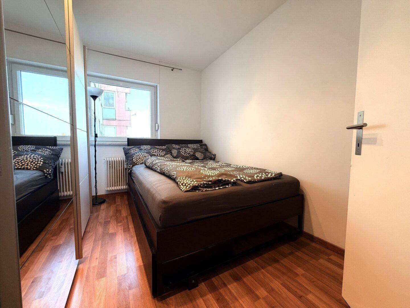 Schlafzimmer mit Doppelbett, Spiegelschrank und Fenster, ausgestattet mit Holzoptikboden.