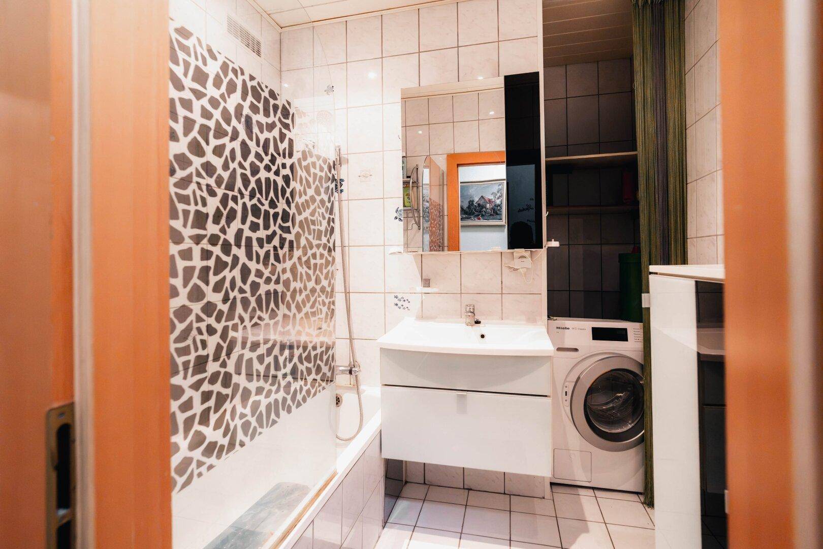 Badezimmer mit Badewanne, Duschfunktion, Waschbecken und Waschmaschine, hell gefliest und praktisch.