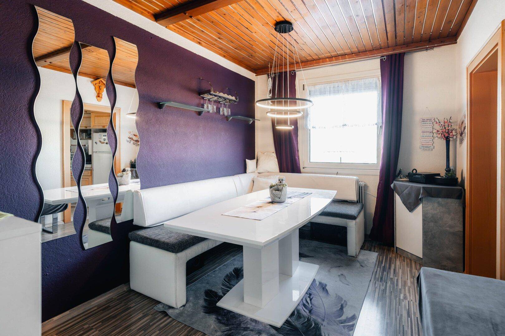 Gemütlicher Essbereich mit Holztisch, Eckbank und auffälliger lila Wandfarbe unter Holzdecke, individuell gestaltet.