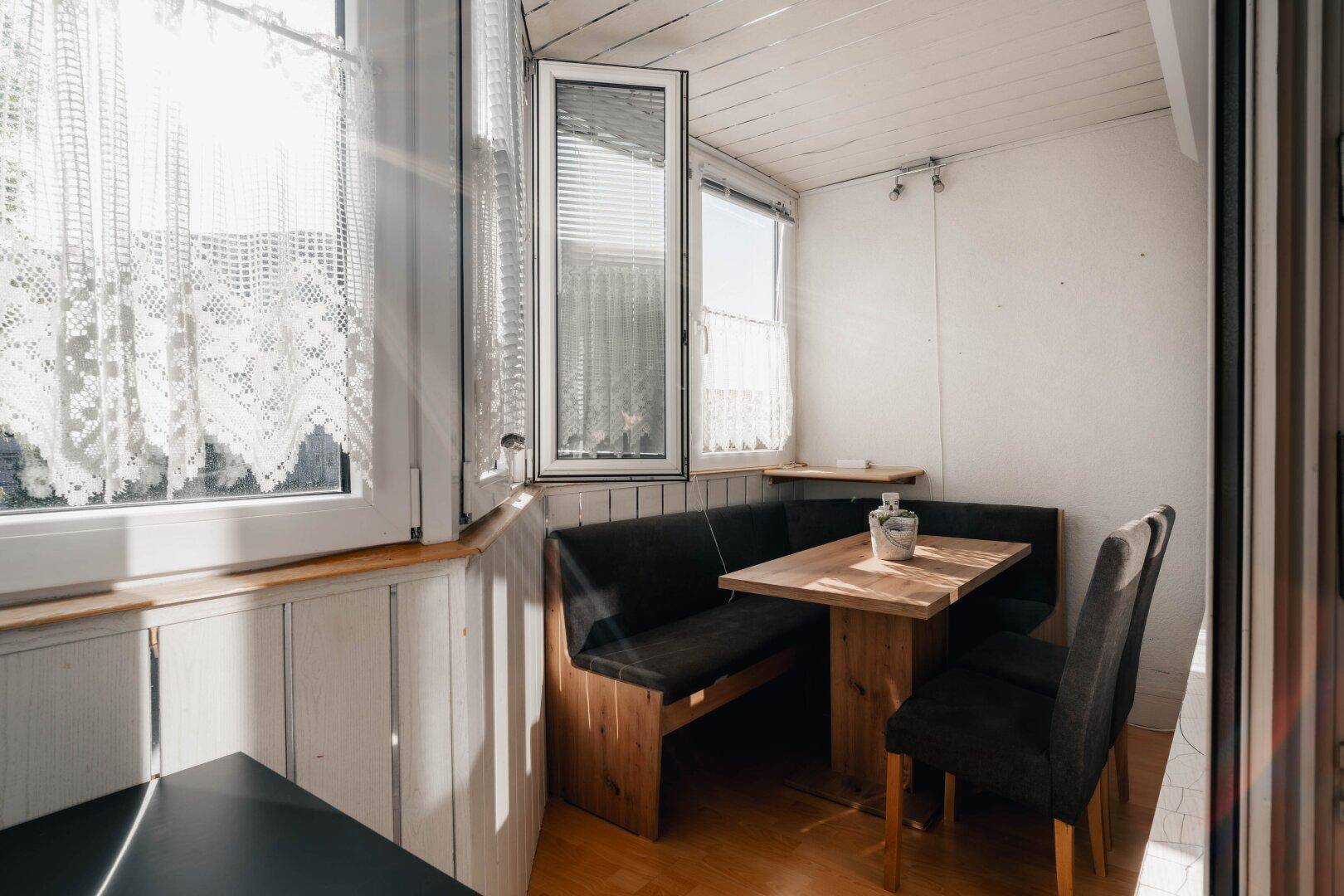 Heller, verglaster Balkon mit Holztisch und Eckbank, perfekt für Mahlzeiten im Freien oder als Leseecke.