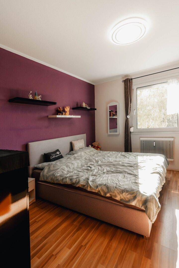 Schlafzimmer mit Doppelbett, lila Akzentwand und Holzboden, sonnig und gemütlich für erholsame Nächte.