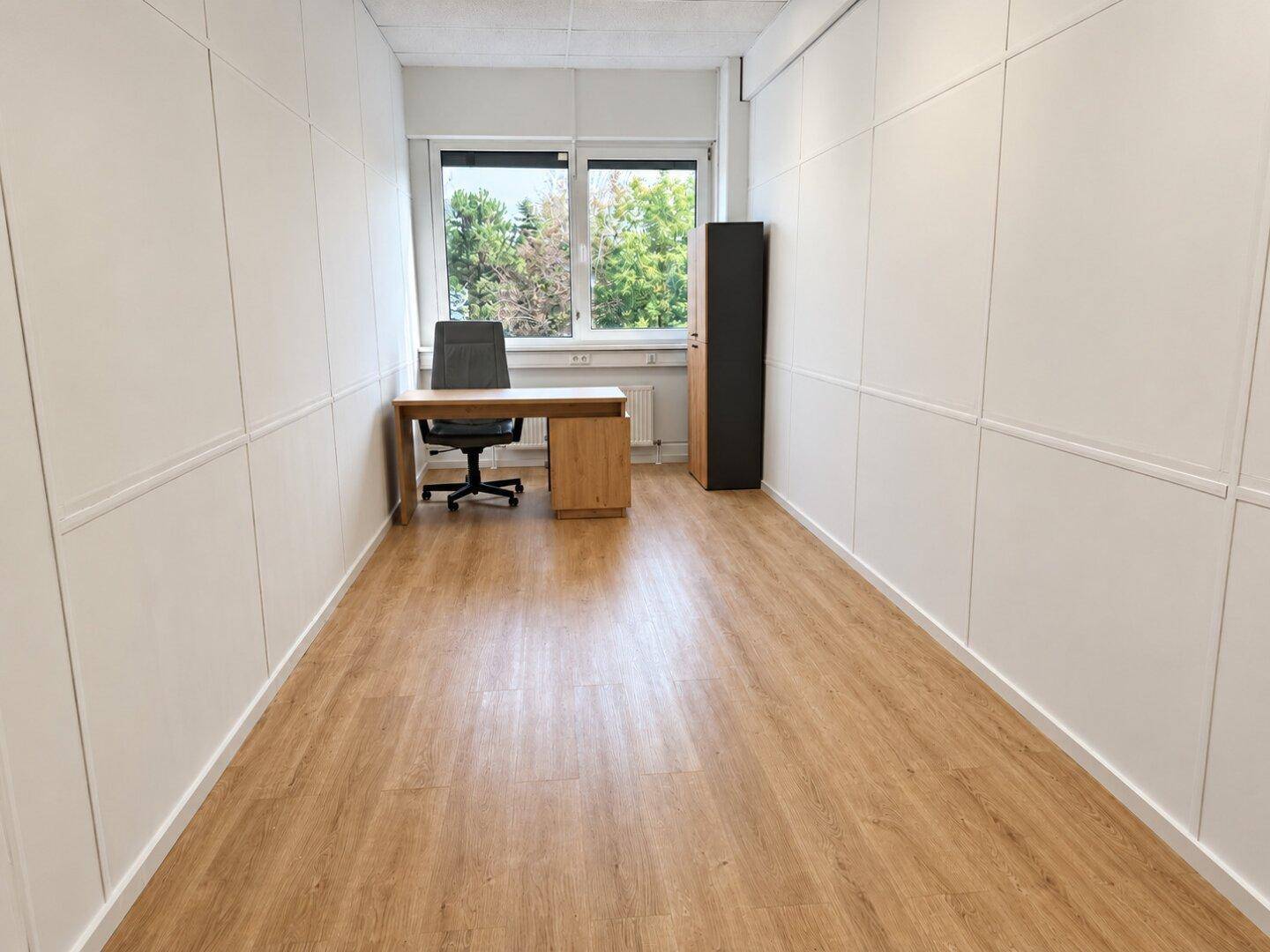 Helles Einzelbüro mit großem Fenster, Holzboden und schlichten weißen Wänden, bereit zur Einrichtung.