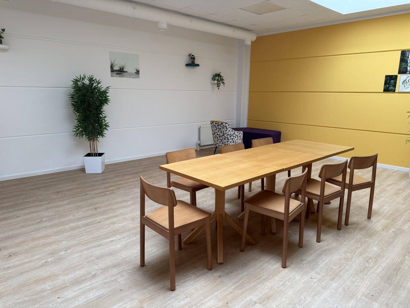 Heller Essbereich oder Besprechungsraum mit Holztisch, Stühlen und gelber Akzentwand, freundlich gestaltet.