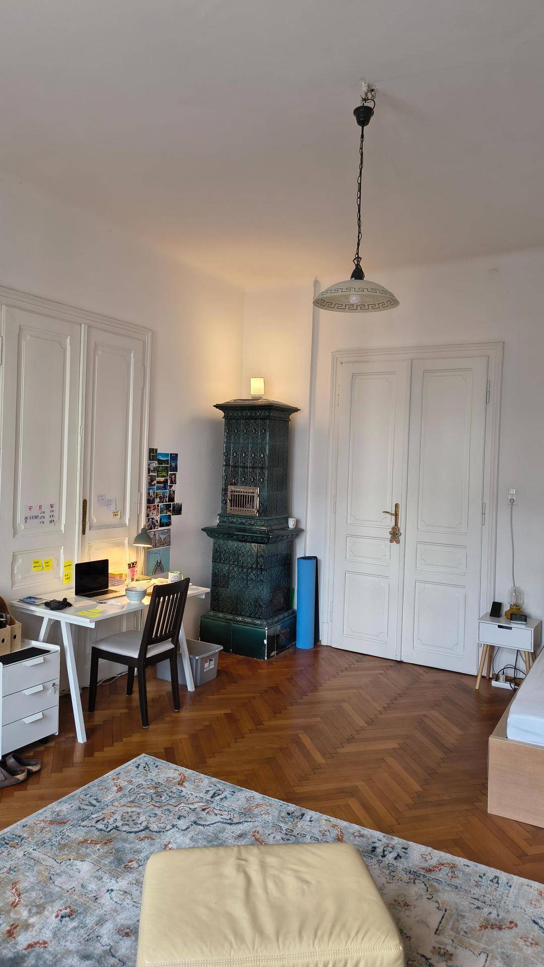 Geräumiges Zimmer mit Fischgrätparkett, einem traditionellen Kachelofen und einem Schreibtischbereich.