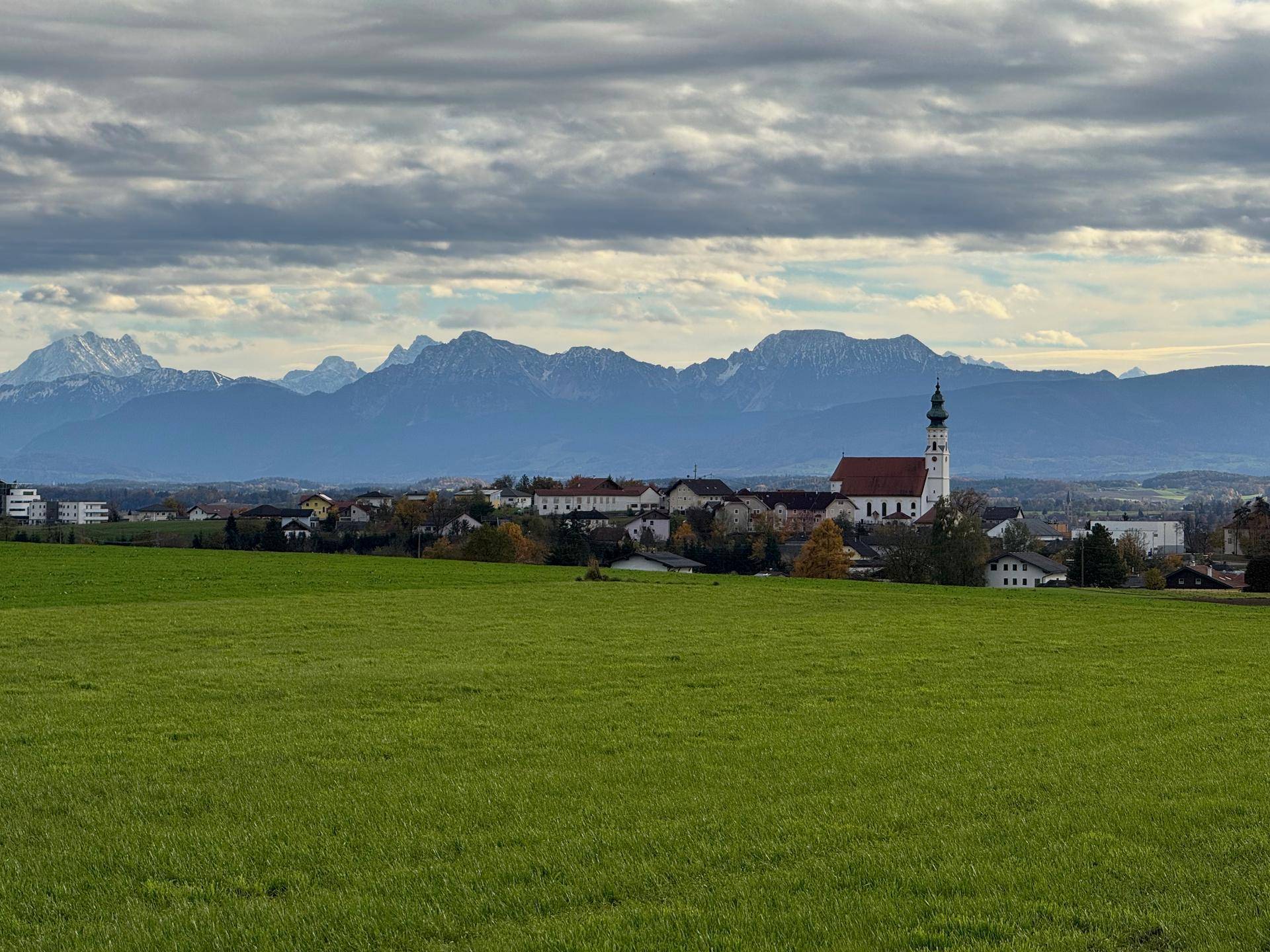 Weitläufige Landschaft mit grünen Feldern, einem Dorf und schneebedeckten Bergen im Hintergrund.