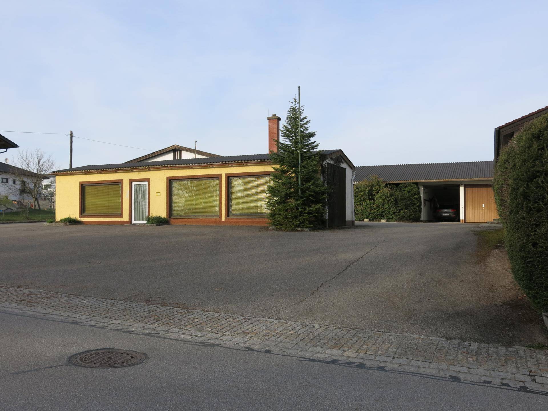 Ansicht des gelben Gebäudes und einer Garage mit großem gepflasterten Bereich und Tannenbaum.