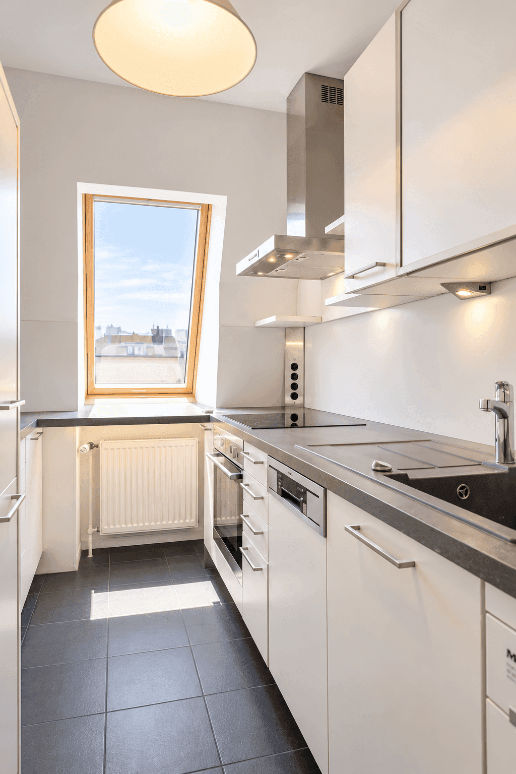 Schmale Küche mit weißen Schränken, dunkler Arbeitsplatte und Dachschrägenfenster.