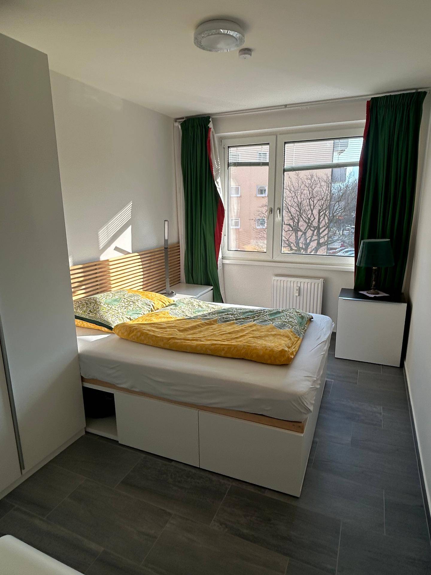 Schlafzimmer mit Doppelbett, Holz-Kopfteil, großem Fenster und dunklen Fliesen.