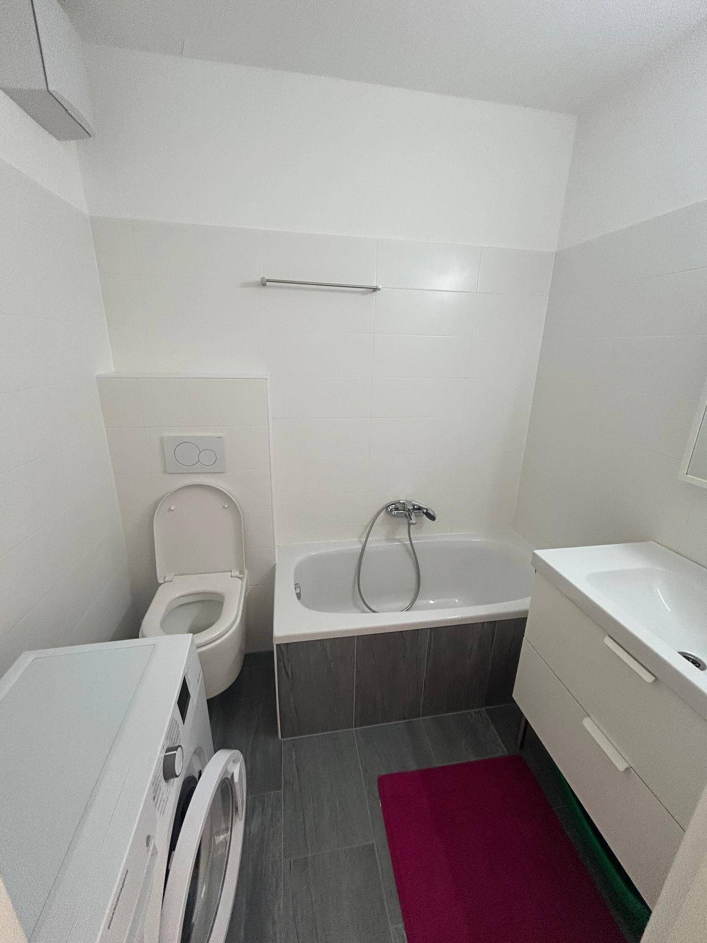 Badezimmer mit Badewanne, wandhängendem WC und Waschmaschine auf Fliesenboden.