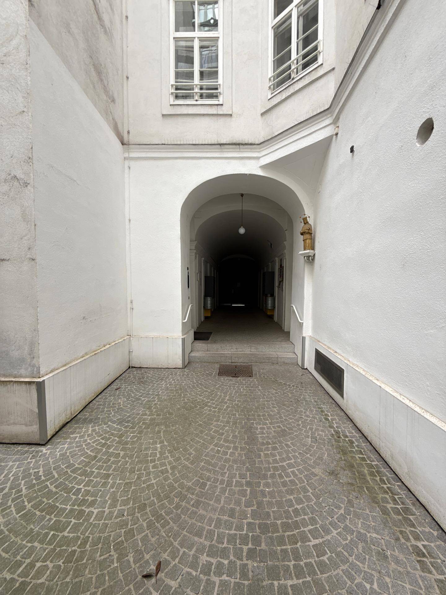 Historischer Innenhof mit Kopfsteinpflaster, der zu einem dunklen Torbogen und weiteren Gebäudeteilen führt.