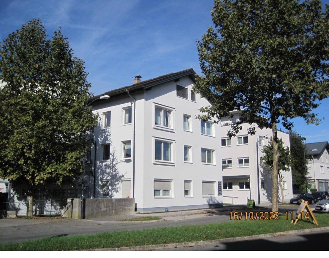 Modernes Wohngebäude mit weißer Fassade und großen Fenstern, umgeben von Bäumen an einem sonnigen Tag.