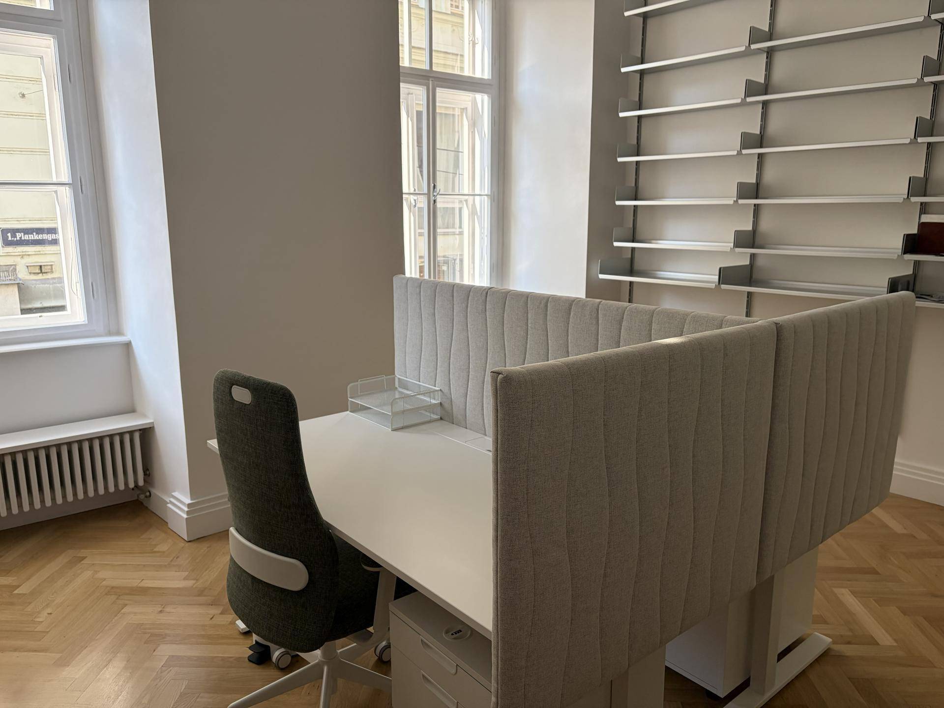 Heller Büroraum mit modernem Schreibtisch, ergonomischem Stuhl und Wandregalen, beleuchtet durch große Fenster.
