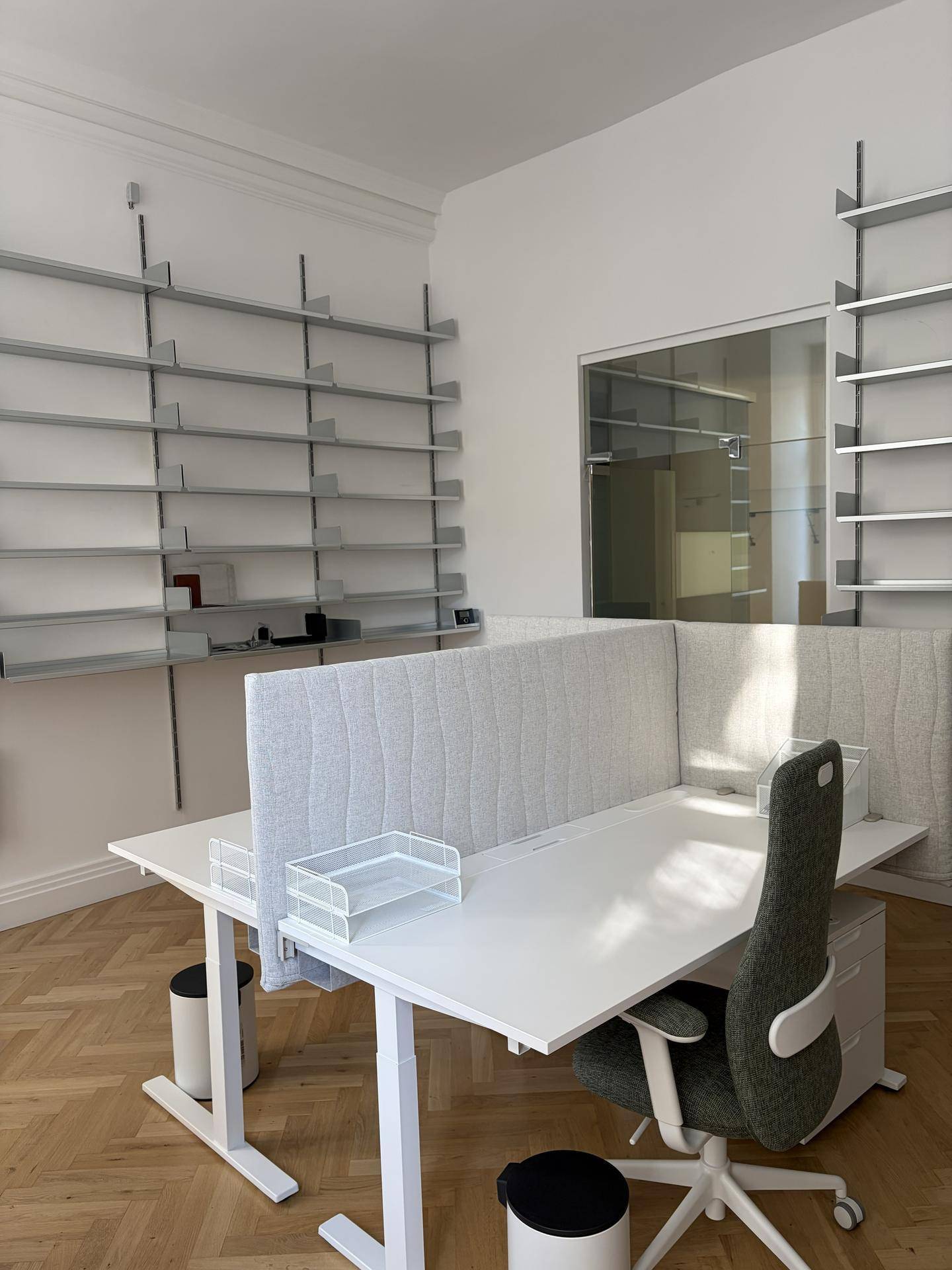 Geräumiges Büro mit zwei Arbeitsplätzen, umfangreichen Wandregalen und einer Glastrennwand im Hintergrund.