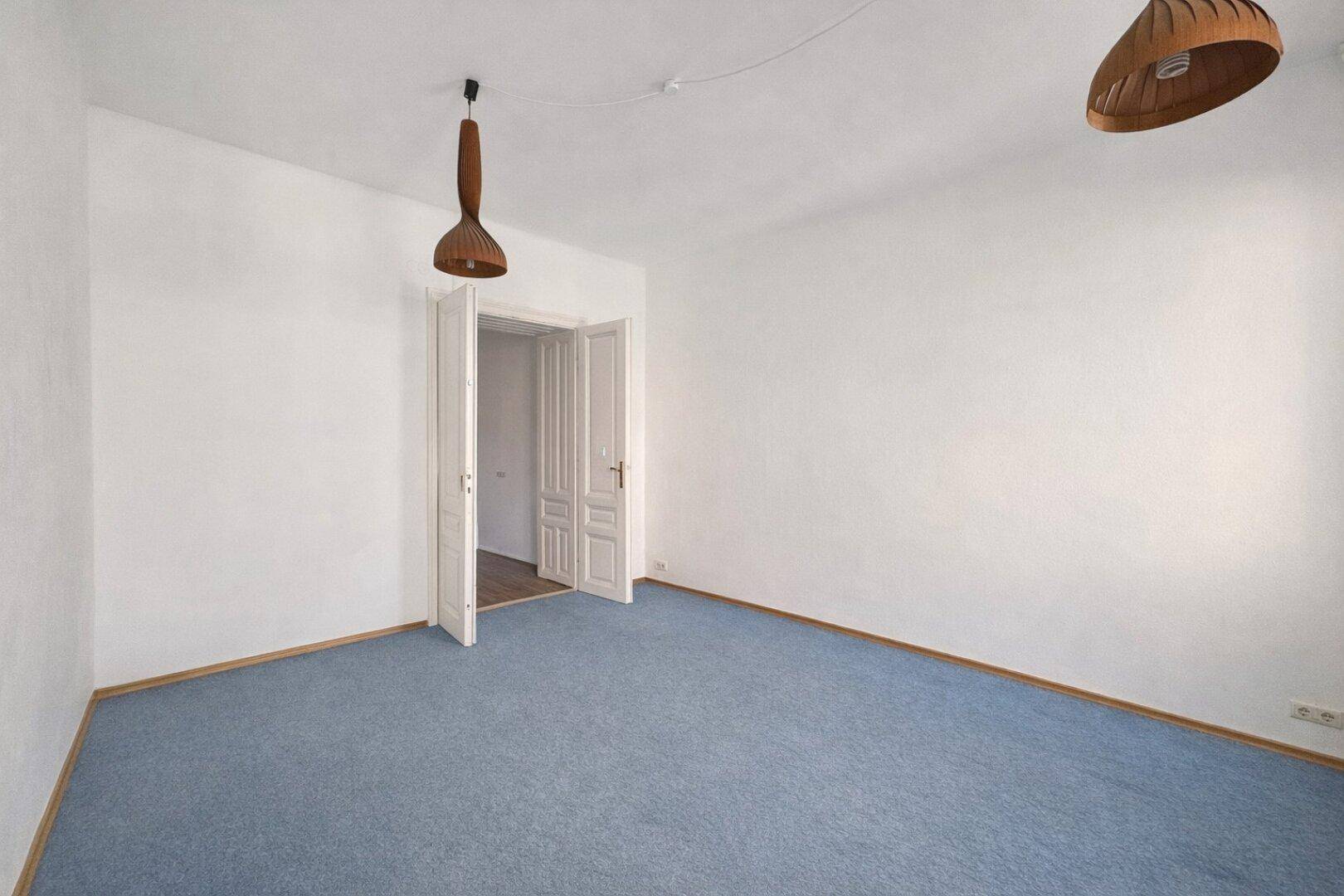 Geräumiger, leerer Wohnbereich mit blauem Teppichboden, weißen Wänden und geöffneten Doppeltüren zu einem weiteren Raum.