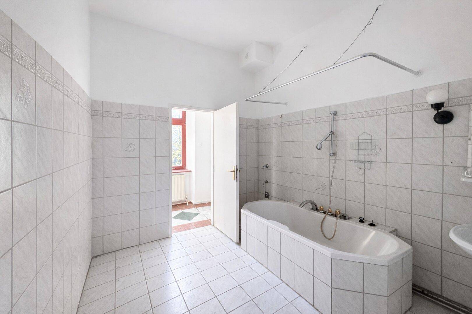 Badezimmer im Stil der 80er Jahre mit weißen Fliesen, einer Eckbadewanne und einem Fenster im Hintergrund.
