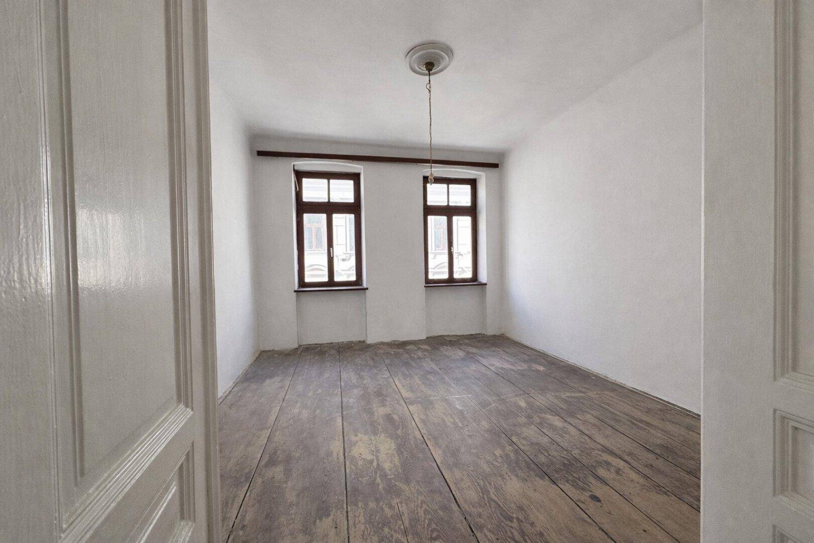 Helles, leeres Zimmer mit Holzdielenboden und zwei Fenstern, frisch gestrichen und bezugsfertig.