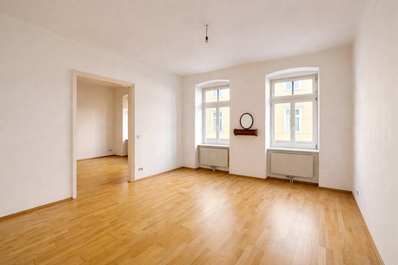 Geräumiges, helles Zimmer mit Holzparkett und Blick in einen weiteren Raum.