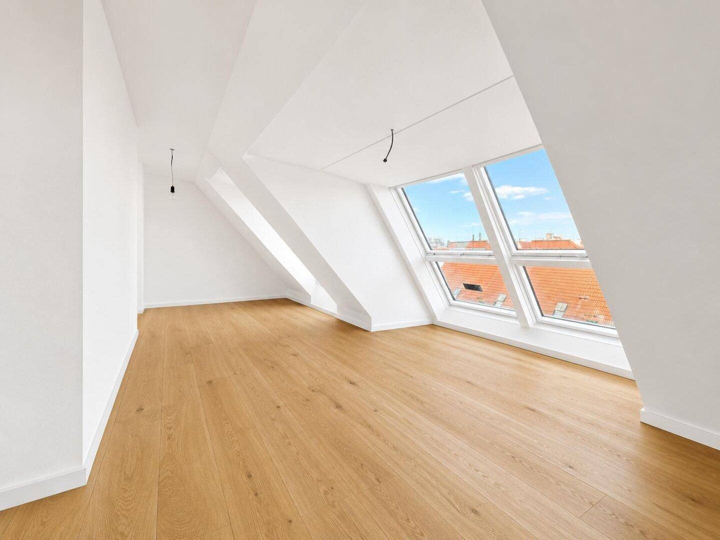 Heller, leerer Raum mit Holzboden und schrägen Wänden, ausgestattet mit großen Dachfenstern.