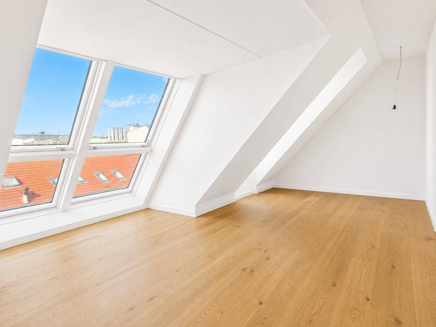 Leerer Raum mit Holzboden, weißen Wänden und großen Dachfenstern, die viel Licht hereinlassen.