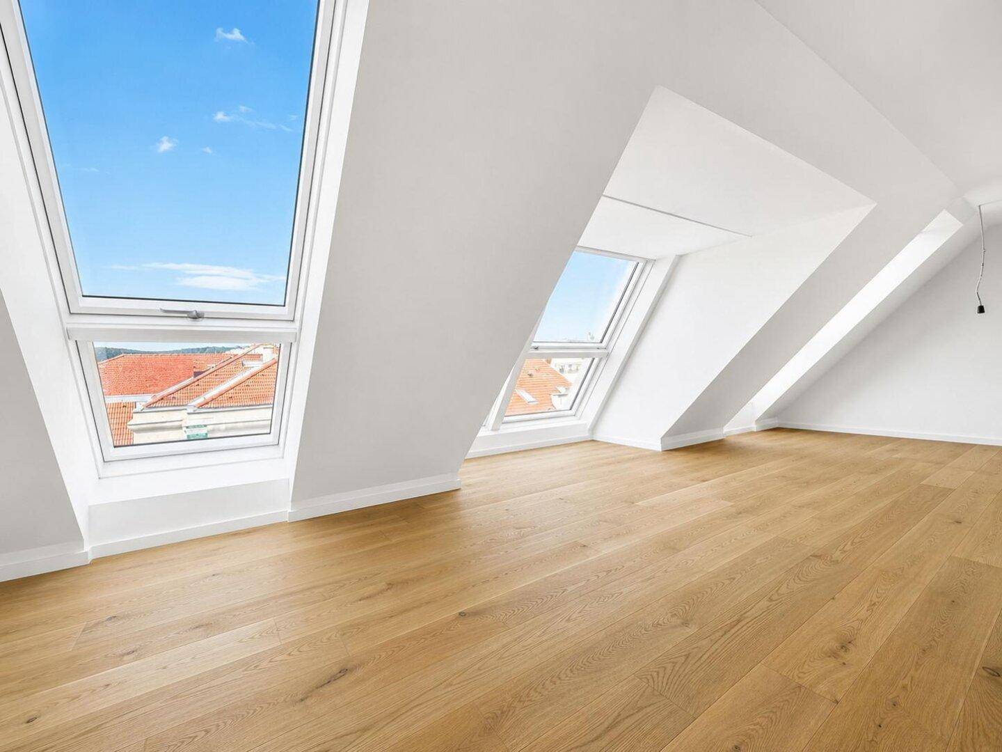 Heller, leerer Raum mit Holzboden und schrägen Wänden, ausgestattet mit großen Dachfenstern.