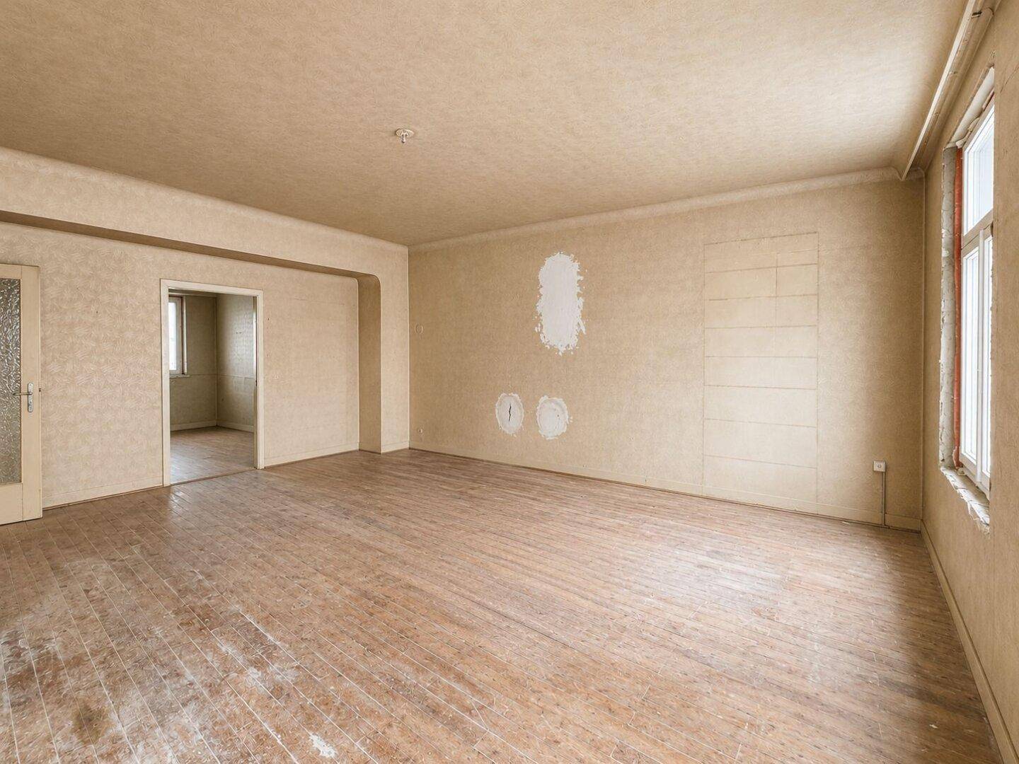 Geräumiges, unrenoviertes Zimmer mit abgenutztem Holzboden und Tapeten, bereit zur Neugestaltung.