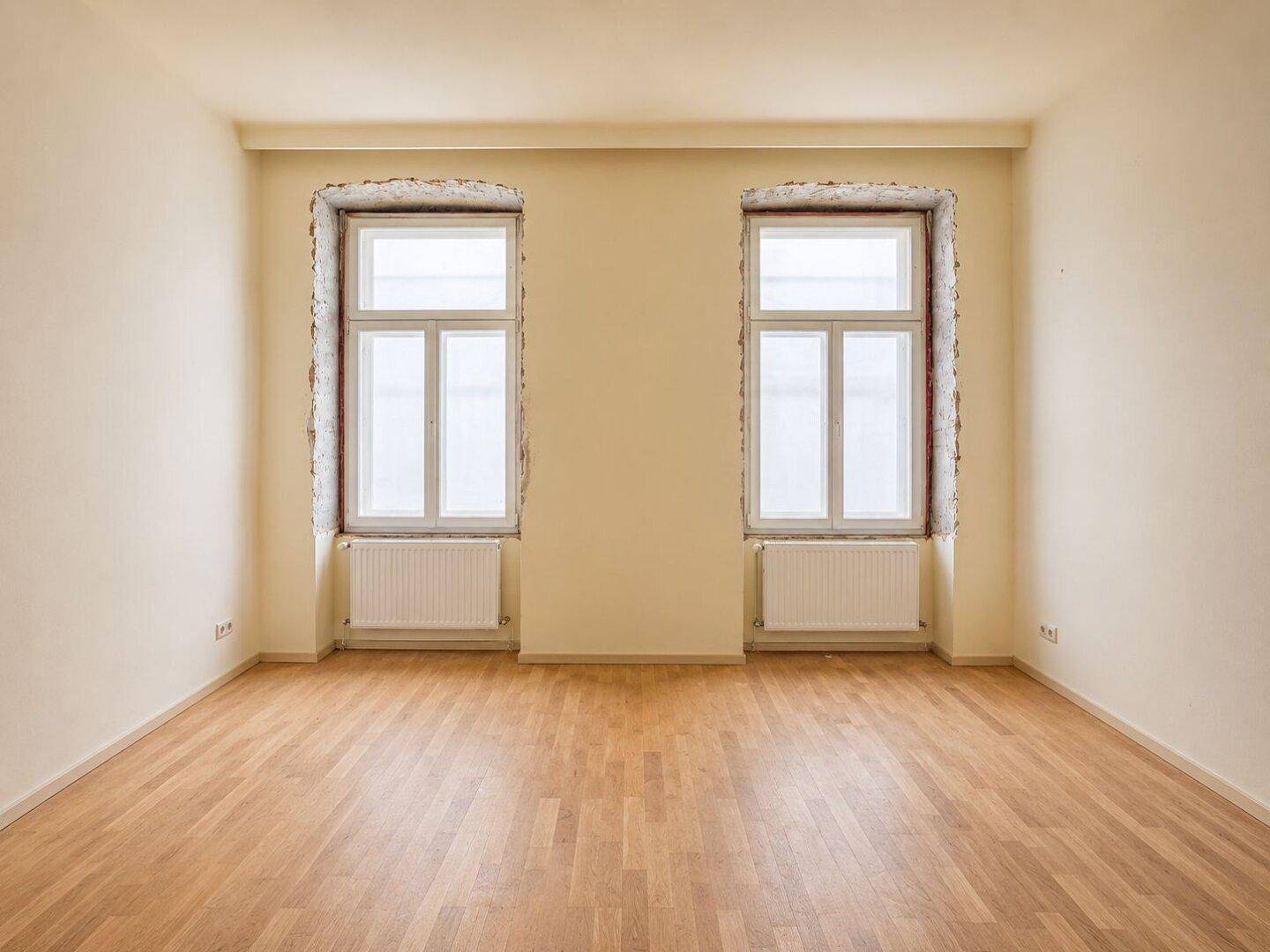 Renoviertes Zimmer mit hellem Parkettboden und zwei großen Fenstern, ideal für Wohnzwecke.