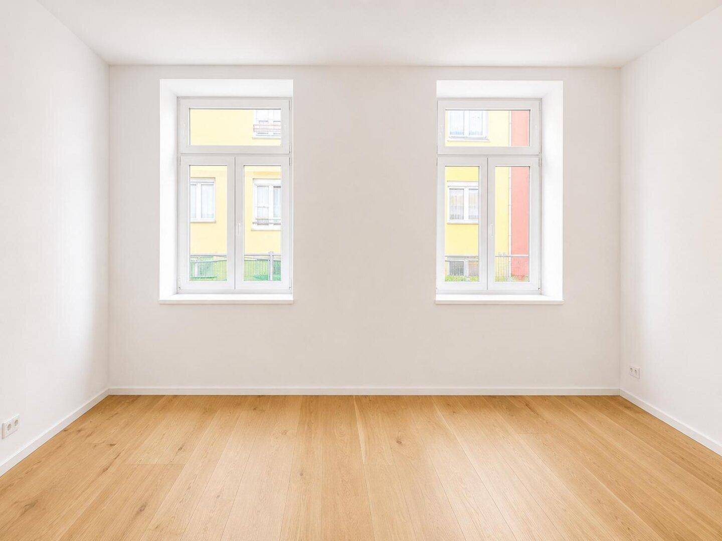 Leeres Zimmer mit hellem Holzparkett und zwei großen Fenstern, die viel Tageslicht hereinlassen.