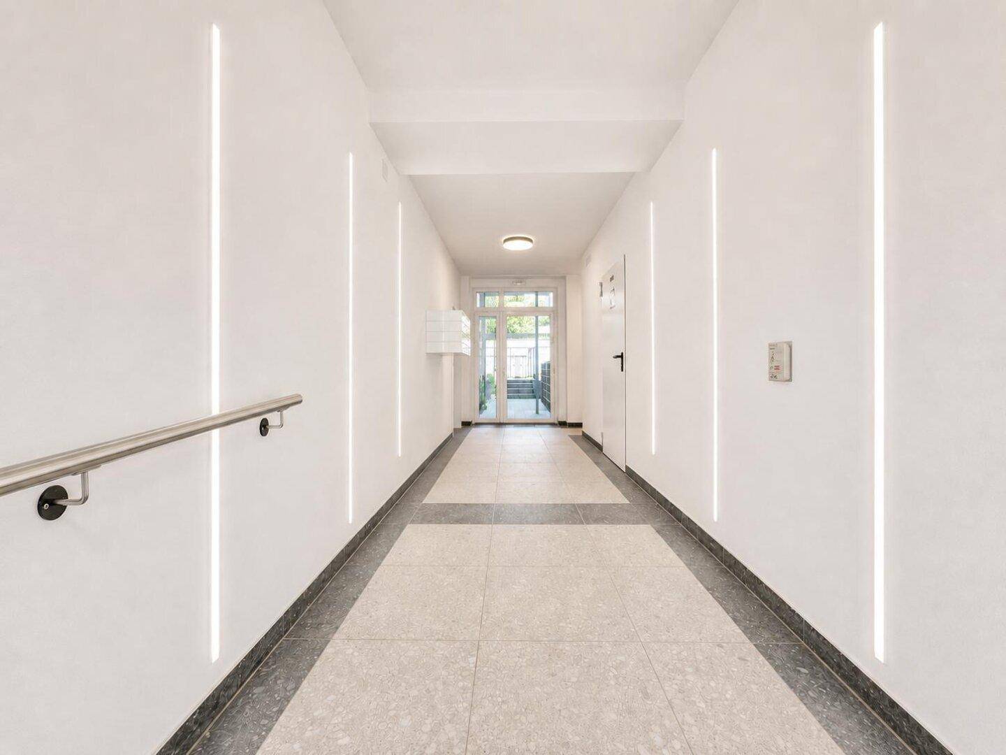 Heller Eingangsbereich mit modernen Lichtleisten, Fliesenboden und einem Handlauf für Barrierefreiheit.