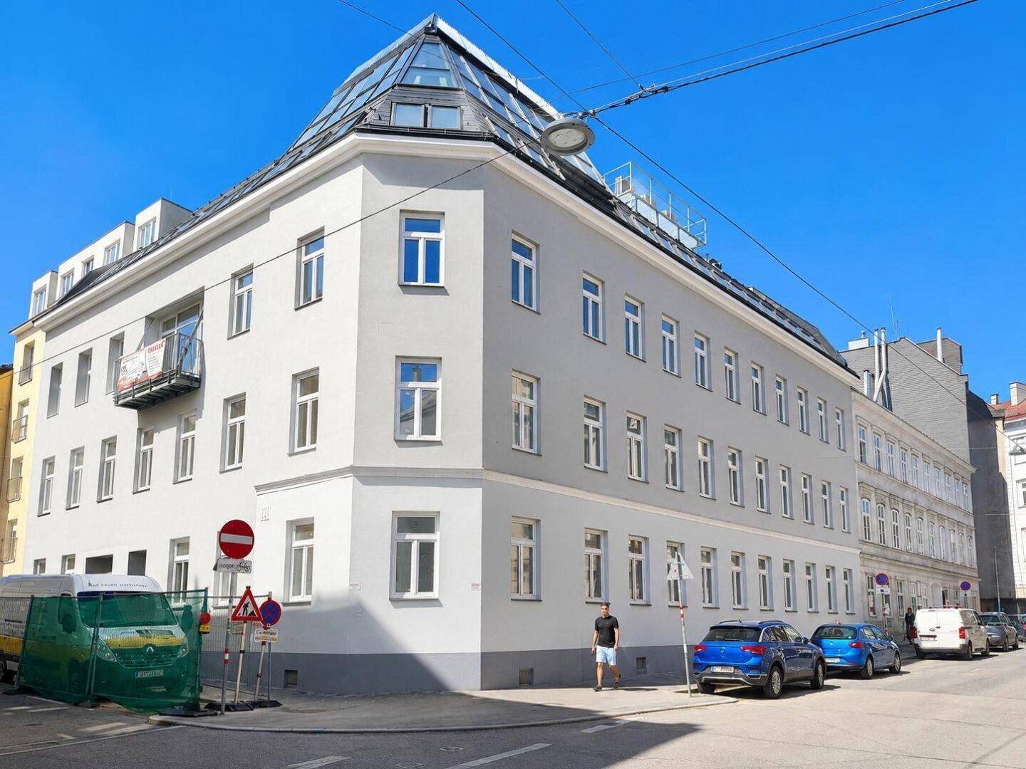 Modernes Eckgebäude mit vielen Fenstern und einem gläsernen Dachaufbau in urbaner Umgebung.