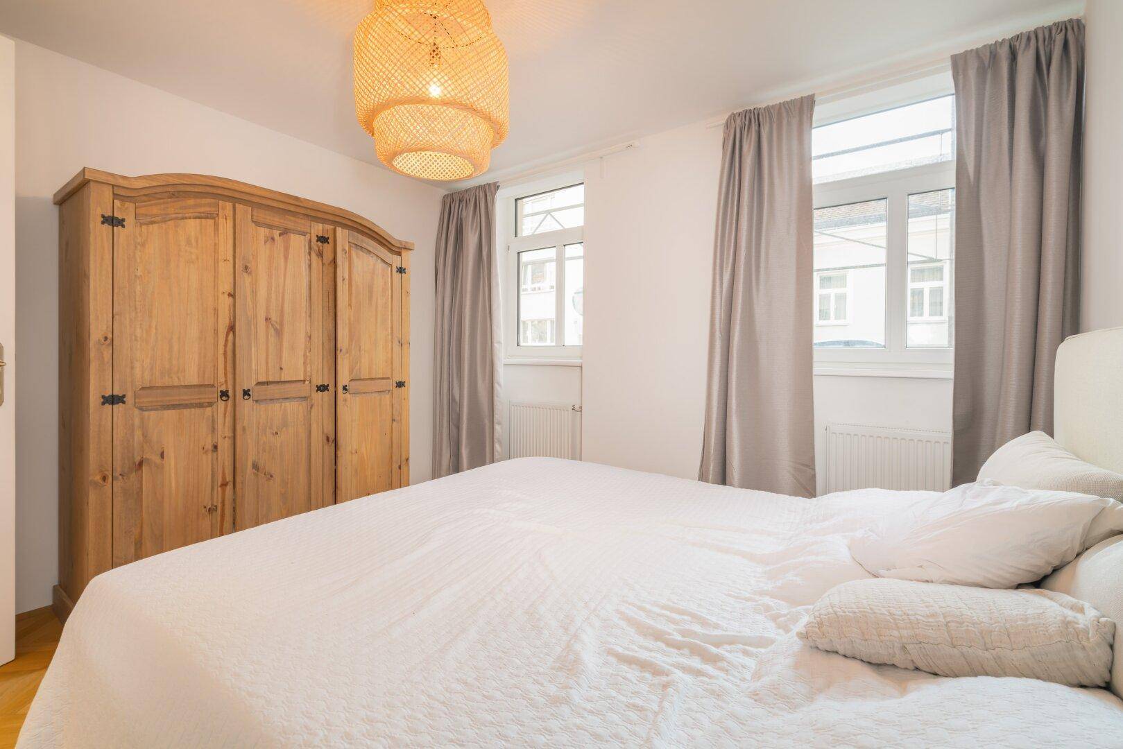 Gemütliches Schlafzimmer mit Holzboden, großem Kleiderschrank und zwei Fenstern mit Vorhängen.