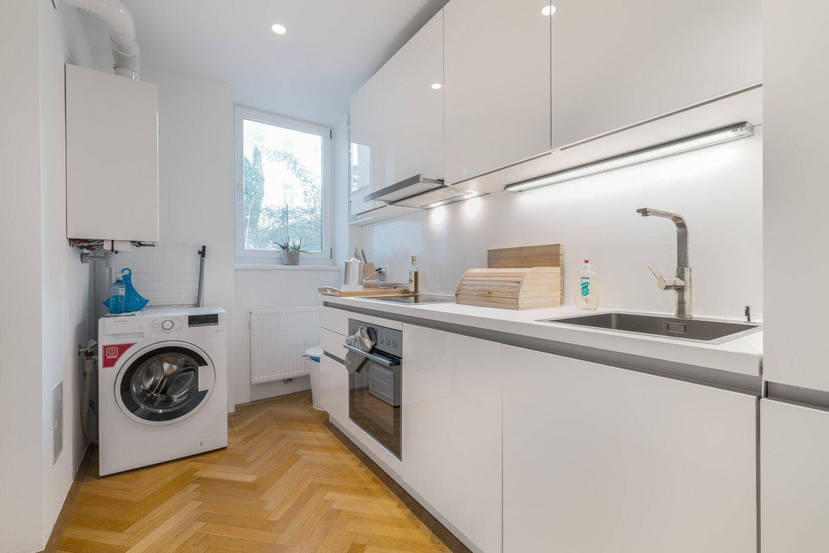 Moderne weiße Küche mit glänzenden Schränken, Waschmaschine und Fenster.