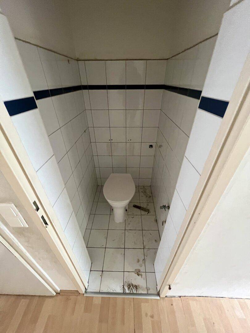 Kleines WC mit weißen Fliesen und blauen Akzentstreifen, in einfachem Zustand.