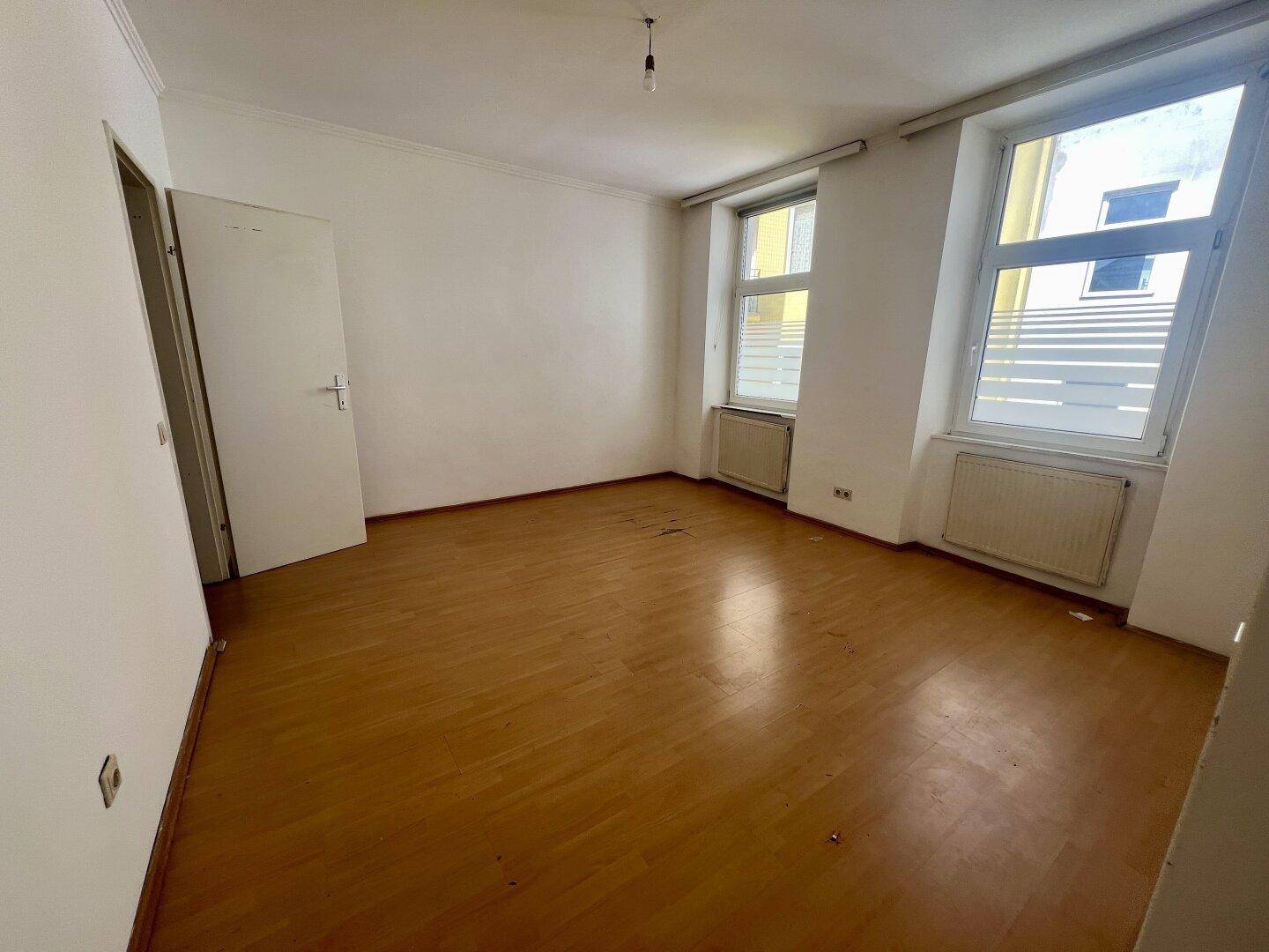 Leerer Raum mit Holzparkettboden und zwei Fenstern, die viel Tageslicht hereinlassen.