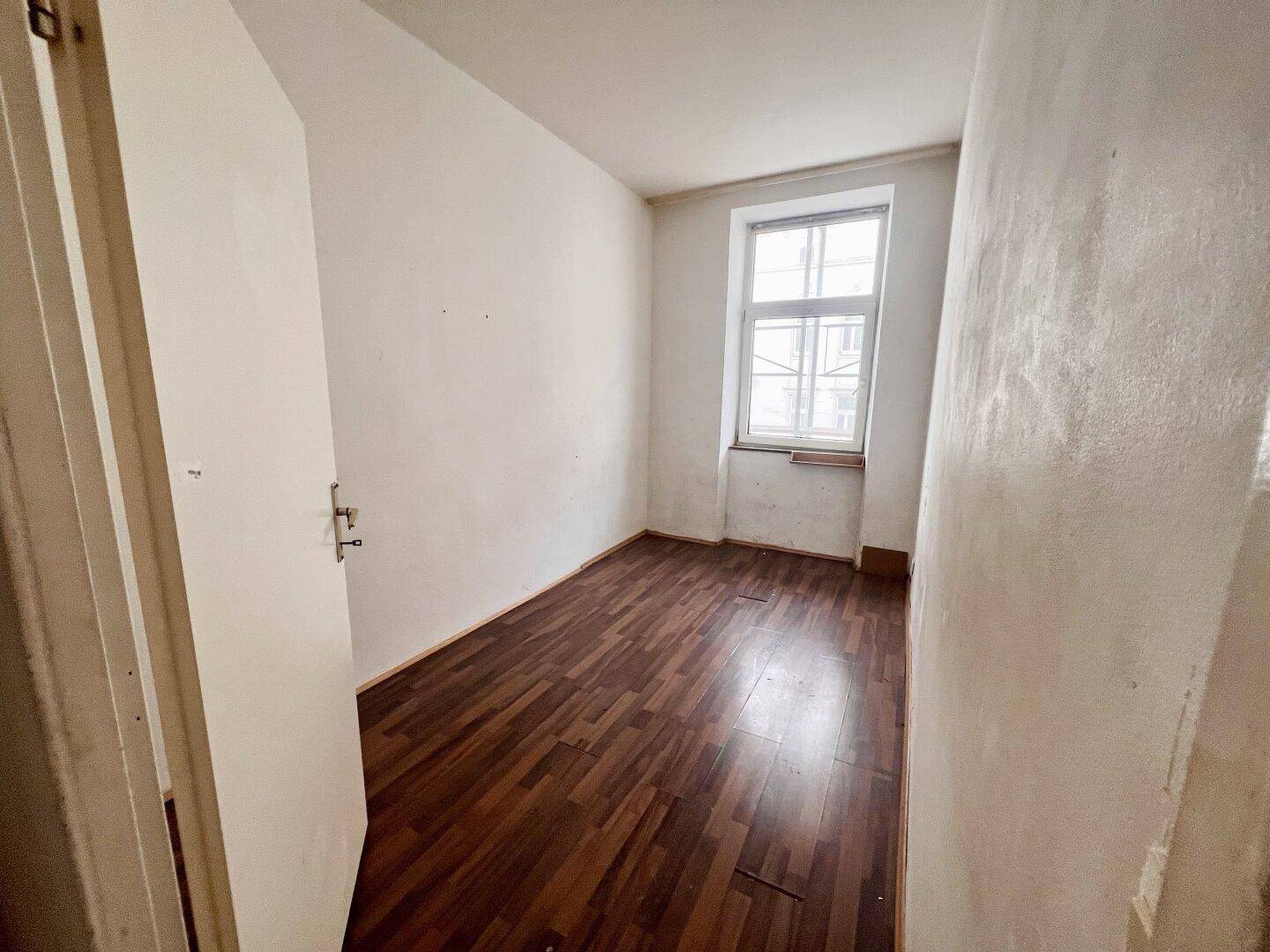 Kleines Zimmer mit dunklem Laminatboden und einem Fenster, ideal als Schlaf- oder Arbeitszimmer.