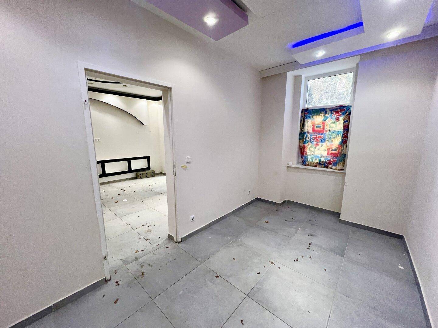 Helles Zimmer mit Fliesenboden, einem Fenster und Blick in einen angrenzenden Raum.