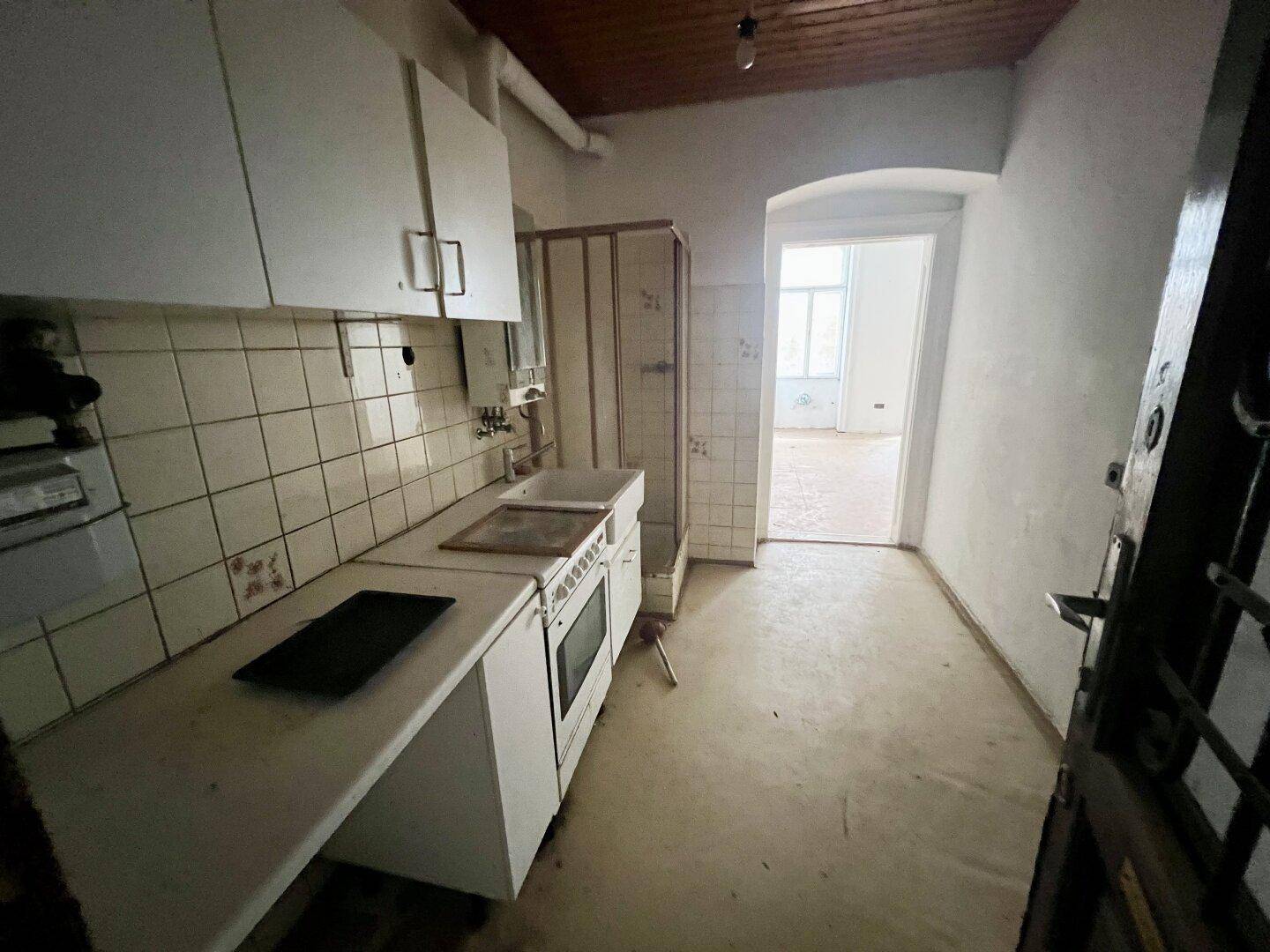 Einfache, ältere Küche mit weißen Schränken, gefliesten Wänden und Blick in einen angrenzenden Raum.