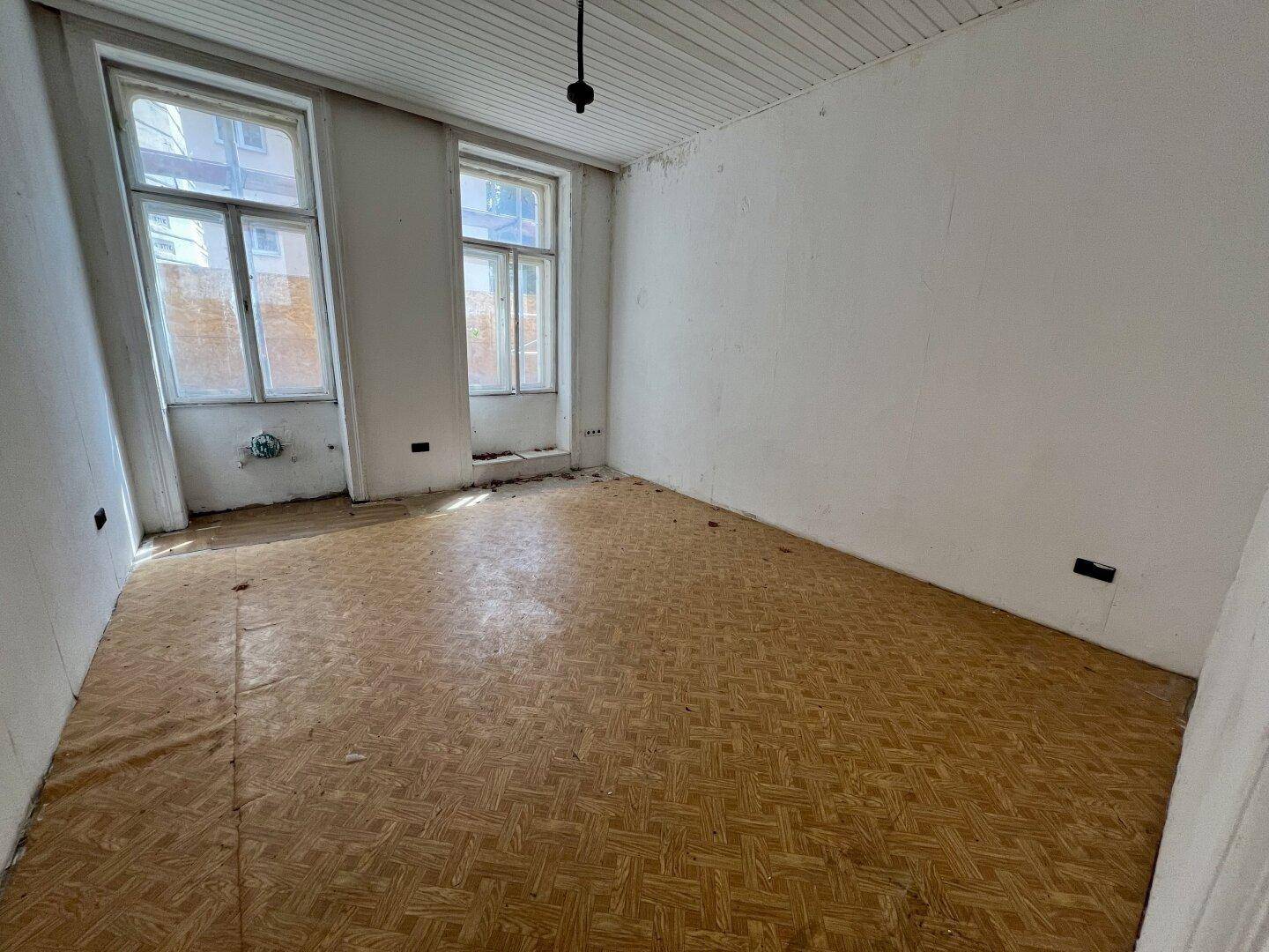 Leerer Raum mit zwei großen Fenstern, Holzboden und einfacher Deckenverkleidung, bereit zur Renovierung.