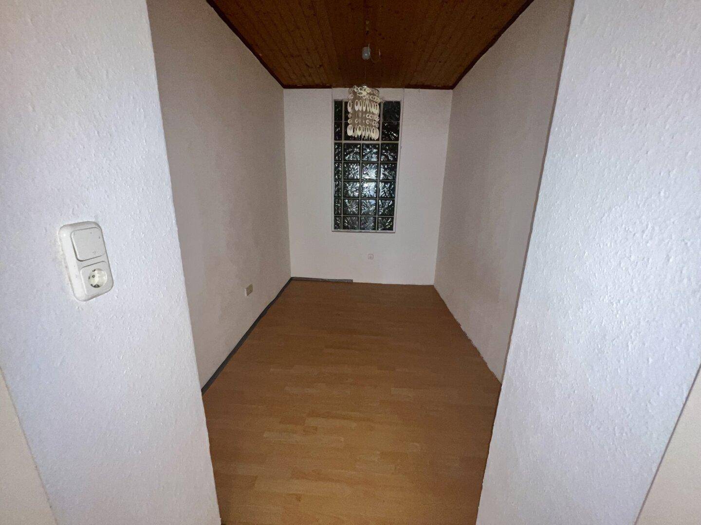 Schmaler Raum mit Holzdecke, Parkettboden und einem Fenster mit Glasbausteinen.