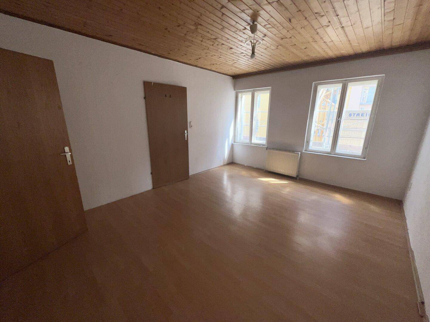 Leeres Zimmer mit Holzbalkendecke, Parkettboden und zwei Fenstern zur Straße.