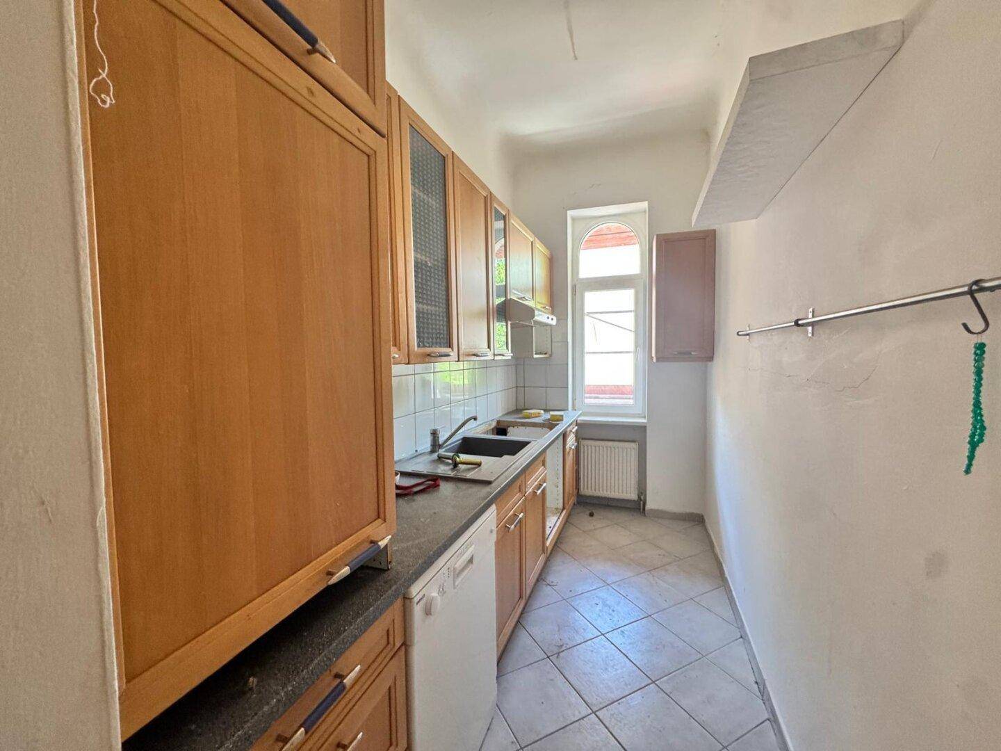 Schmale Küche mit Holzschränken, Fliesenboden und Fenster, funktional eingerichtet.