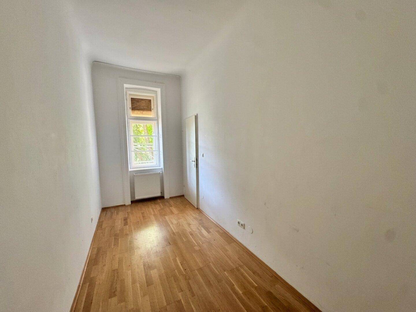 Helles, leeres Zimmer mit Holzboden und einem Fenster, das Tageslicht spendet.