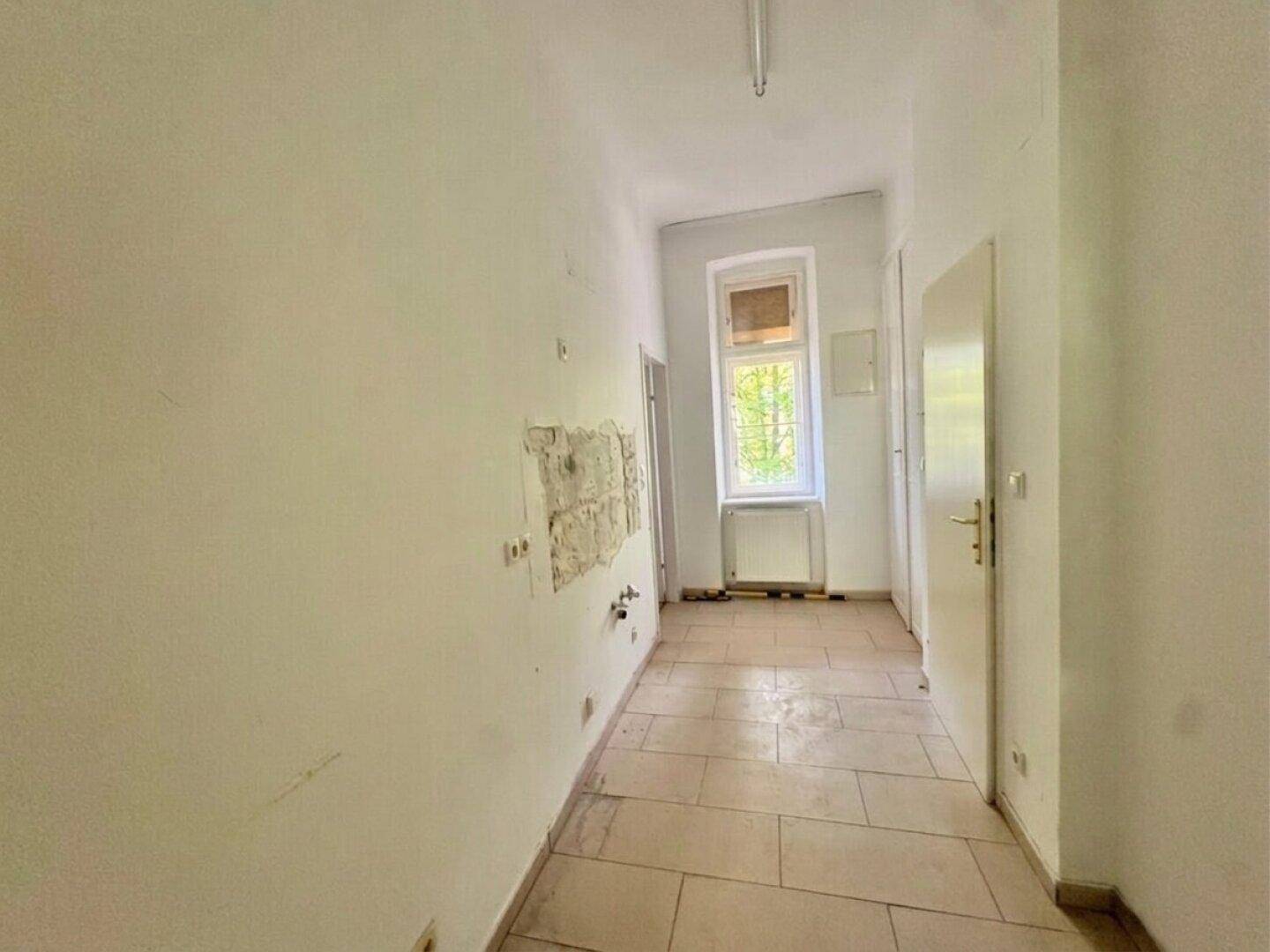 Langer Flur mit Fliesenboden, einem Fenster und sichtbaren Wandanschlüssen.