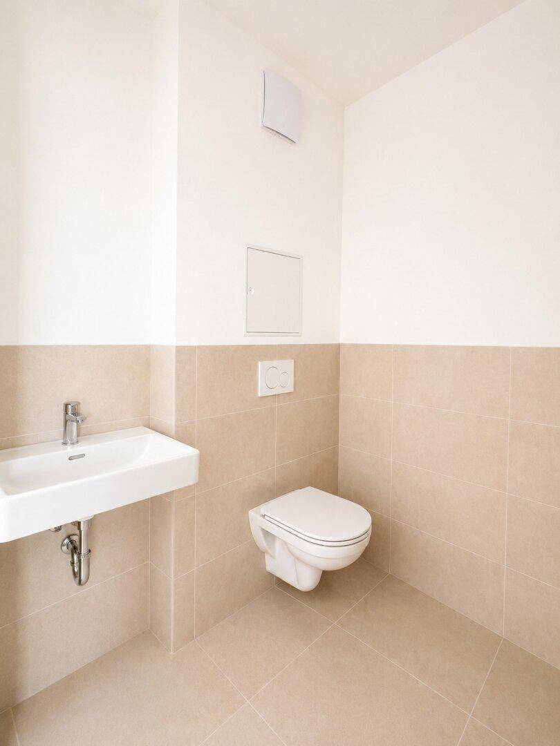 Funktionelles WC mit wandhängender Toilette, Einzelwaschbecken und beigefarbenen Wand- und Bodenfliesen.